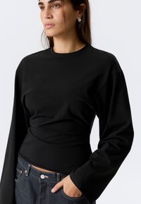 Haut noir à manches longues avec un col rond et une taille ajustée, fabriqué en tissu lisse. Associé à un denim foncé, avec passants de ceinture.