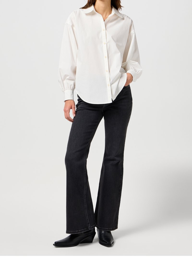 Chemise blanche à manches longues avec boutons, coupe décontractée, ourlet arrondi, associée à un jean noir évasé et des bottines noires à bout pointu.