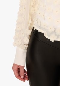 White blouse with 3D floral appliqués and a hemline, paired with high-waisted black faux leather leggings. Long, sheer, textured sleeves.