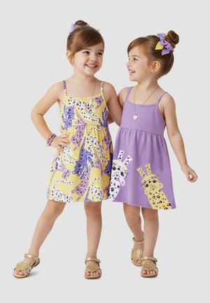 Dwie młode dziewczynki uśmiechają się, noszą letnie sukienki bez rękawów z nadrukiem zwierząt, pasujące złote sandały i mają wiązane kokardki we włosach, stoją obok siebie.