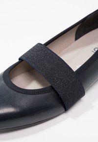 Chaussure plate Mary Jane noire avec tige en cuir texturé et large bride élastique scintillante sur le dessus, semelle intérieure beige rembourrée visible.