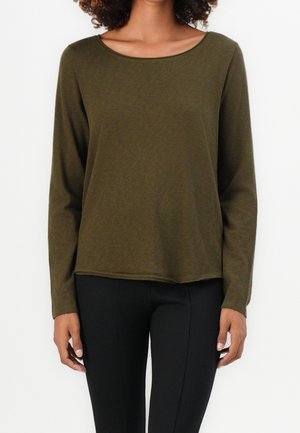 Frau trägt ein olivgrünes Langarmoberteil mit Rundhalsausschnitt und schwarze eng anliegende Hose, steht vor einem schlichten weißen Hintergrund.