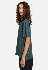 T-shirt in cotone verde scuro con vestibilità rilassata, maniche corte e dettagli di cucitura minimi. Il modello sta in piedi di lato, con le braccia rilassate.