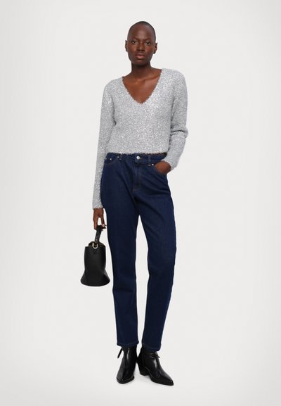 Jersey plateado, brillante, de manga larga con escote en V, combinado con jeans rectos de un azul oscuro. Sostiene un pequeño bolso negro y lleva botas de tobillo con punta.