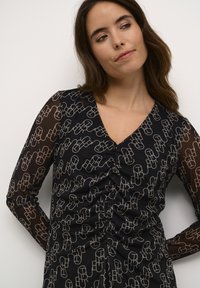 Črna obleka z dolgimi prozornimi rokavi, ki ima geometrijski vzorec v bež barvi in spredaj nabran detajl za teksturo in prileganje.