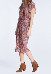Blumiges Midikleid in Rosa-, Orange- und Lilatönen. Volantsärmel, taillierter Schnitt und fließende Silhouette. Kombiniert mit braunen Knöchelschuhen.