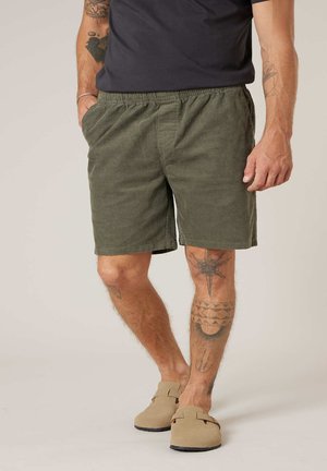 Homme portant un short en velours côtelé vert olive, un t-shirt noir, des chaussures beige sans lacets, avec des tatouages visibles sur les bras et les jambes.