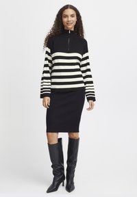 Pull à col roulé noir et crème rayé avec fermeture éclair, associé à une jupe crayon noire côtelée et des bottes noires jusqu'aux genoux.