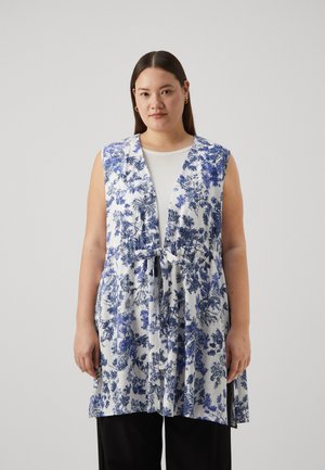 Blomstervest i hvit med blå mønstre, med knytelukking, åpen front og uten ermer. Kombinert med en hvit topp og svarte bukser.