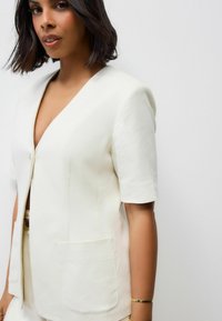 Bílý krátký rukávový blazer s výstřihem do V, s jedním knoflíkovým zapínáním a přední kapsou. Hladký texturní materiál, moderní střih.