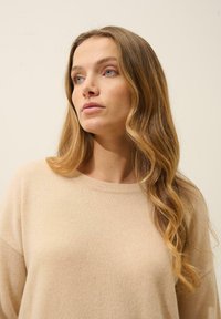Beige pletený svetr s kulatým výstřihem a dlouhými rukávy, který má měkkou texturu a volný střih. Vlasy jsou vlnité a po ramena.