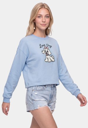 Ung kvinde med langt blondt hår iført en blå sweatshirt med en tegneseriehund og teksten "Love Dog" kombineret med slidte denimshorts.