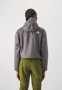 Szara krótka kurtka z kapturem, z logo na plecach. Połączona z zielonymi spodniami, prezentującymi elegancki, wodoodporny design.