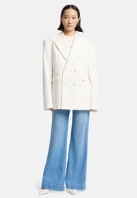 Doppelreiher weißer Blazer mit goldenen Knöpfen, oversized Schnitt, kombiniert mit hellblauen Schlagjeans und weißen spitzen High Heels.