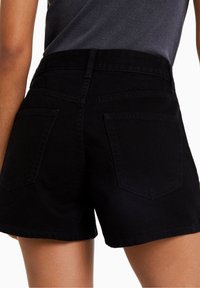 Kvinna i svarta denimshorts och en mörkgrå topp som är instoppad, sedd bakifrån mot en vit bakgrund.