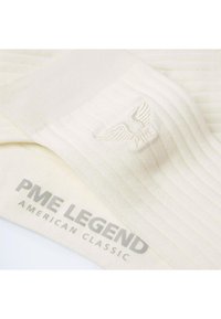 Chaussettes blanches à côtes avec le logo PME Legend et un emblème ailé brodé sur le dessus, pliées sur un emballage de marque assorti.