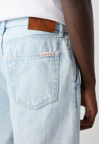 Person wearing light blue denim jeans with Calvin Klein labels on the back pocket and waistband, standing with one arm relaxed.