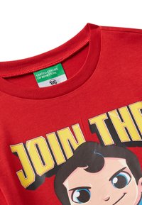 Červené tričko s kulatým výstřihem, žlutý nápis "PŘIDEJTE SE K", a obličej karikatury s velkýma modrýma očima a černými vlasy.