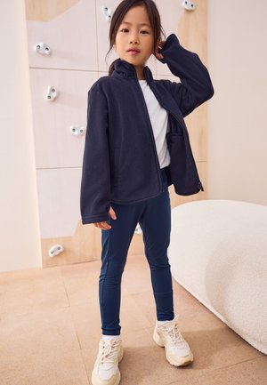 Fiatal lány sötétkék dzsekiben, fehér ingben, kék nadrágban és sportcipőben, beltéren áll a falmászó fal közelében, bézs színű padlón.