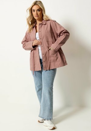 Lyshåret kvinde iført en støvet lyserød knappet jakke over en hvid top, lyseblå jeans og hvide sneakers, stående foran en ensfarvet baggrund.