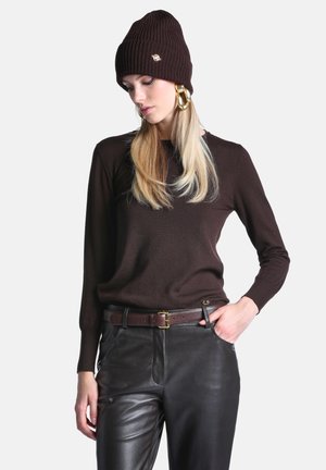 Femme portant un bonnet en maille marron foncé, un pull assorti, un pantalon en cuir noir, une ceinture marron et de grandes boucles d'oreilles créoles dorées sur un fond uni.