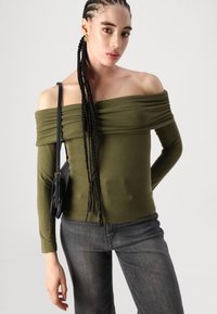 Oliven grønn topp med avtagende skulder, med samlet tekstur, lange ermer og en tettsittende design, kombinert med mørkegrå jeans.