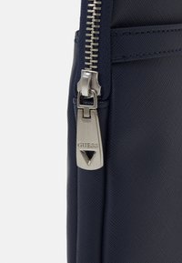 Borsa texture blu navy con dettaglio della cerniera, dotata di un tag di metallo argentato con il logo "GUESS" e un accento triangolare.