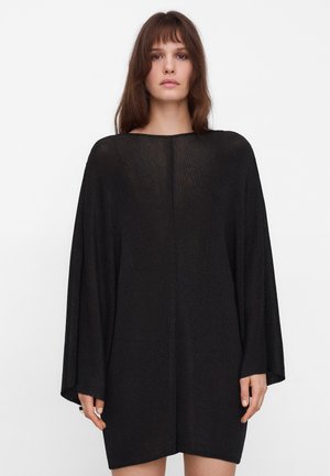 Vrouw met bruin haar, gekleed in een losvallende, zwarte tuniekjurk met lange mouwen, tegen een effen witte achtergrond.