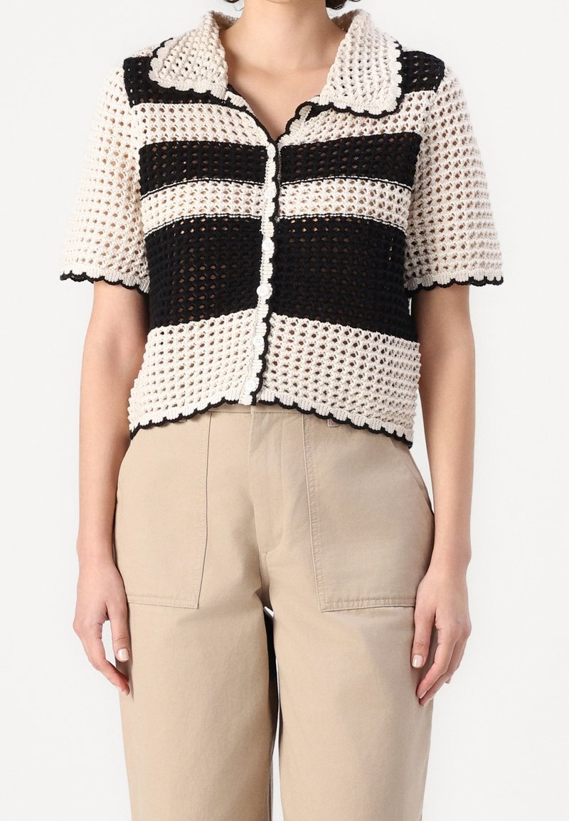 Femme portant un haut boutonné en crochet noir et blanc à manches courtes avec des bords festonnés et un pantalon taille haute beige.