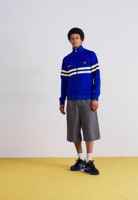 Mėlyna užtrauktuku uždaroma striukė su baltais ir geltonais dryžiais, pilkos šortai ir juodi sportiniai bateliai su mėlynomis akcentais ant geltono grindų.