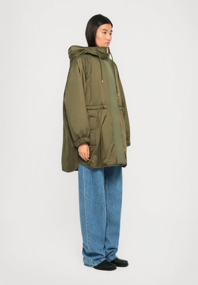 Woman wearing an oversized olive green puffer jacket with hood, wide-leg blue jeans, and black shoes, standing in a neutral background.