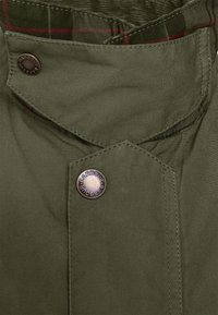 Zielona bawełniana tkanina z podszewką w kratę, z okrągłym metalowym guzikiem z wygrawerowanym napisem "Barbour". Widoczne szwy wzdłuż krawędzi.