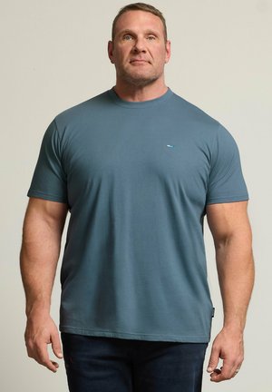 Homme à la carrure musclée portant un t-shirt bleu uni à manches courtes et un pantalon foncé, debout devant un fond neutre.