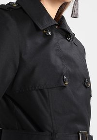 Black trench coat with a classic design, featuring large buttons, a belt, and a structured collar. Smooth fabric with a polished finish.