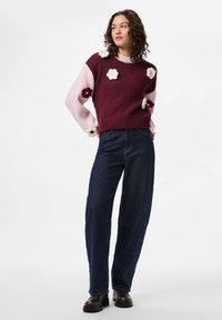 Πλεκτό πουλόβερ σε μπορντό και ροζ χρώμα με λουλουδένιες εφαρμογές, στρογγυλή λαιμόκοψη, ριμπ τελείωμα, συνδυασμένο με σκούρο μπλε παντελόνι φαρδιάς γραμμής και μαύρες μπότες.