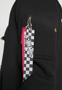 Manche de veste noire avec poche zippée, comprenant deux porte-clés : un rouge avec texte blanc, l'autre à carreaux noir et blanc avec un logo Hot Wheels.