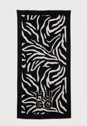 Melnā dvielī ar baltu zebras svītru rakstu un "HUGO" logotipu biezās burtiem netālu no apakšējā centra.