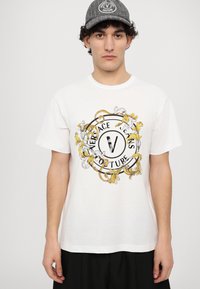 Balta medvilninė marškinėlių su apvaliu grafiniu elementu, kuriame užrašyta "Versace Jeans Couture", papuošta geltonais ir juodais gėlių motyvais.