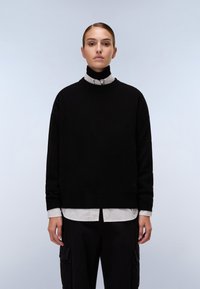 Maglione nero a maglia con scollo rotondo e polsini a coste, indossato sopra una camicia a righe grigio chiaro e bianca con colletto. Pantaloni neri con tasche.