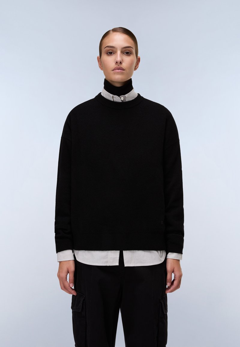 Maglione nero a maglia con scollo rotondo e polsini a coste, indossato sopra una camicia a righe grigio chiaro e bianca con colletto. Pantaloni neri con tasche.