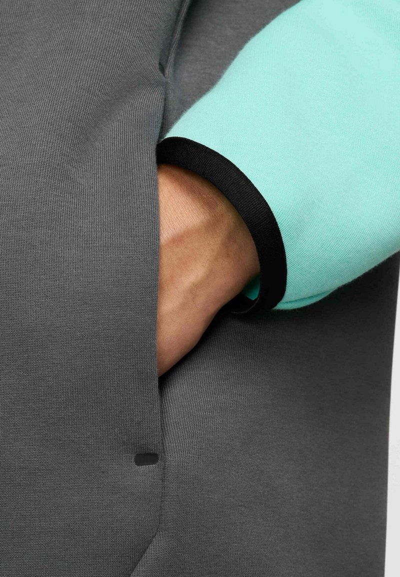 Prenda gris de manga larga con un acento turquesa en el puño, con un ribete negro. Se ve una mano en un bolsillo lateral. Textura suave.