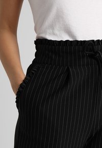 Pantalons noirs à rayures avec une taille élastique à volants, des poches latérales et un tissu doux texturé, présentant un détail à cordon de serrage.