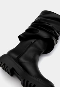 Bota alta negra con diseño holgado, hecha de cuero suave. Presenta un cierre lateral y una suela de goma gruesa con un agarre profundo.