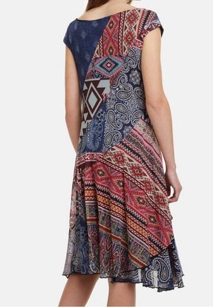 Žena v šatech délky po kolena s nášivkami a kombinací geometrických a paisley vzorů v odstínech červené, modré a růžové, zobrazená zezadu.