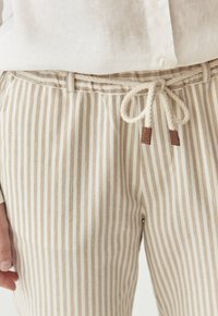 Pantalones beige a rayas con cintura ajustable con cordón y detalles en cuero marrón. Hechos de una tela texturizada, con rayas verticales en tonos claros y oscuros.