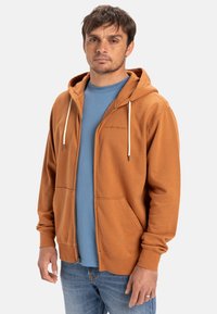 Orange zip-up hoodie med dragsko, framfickor och avslappnad passform, bärs över en blå skjorta, ihop med ljusblå jeans.