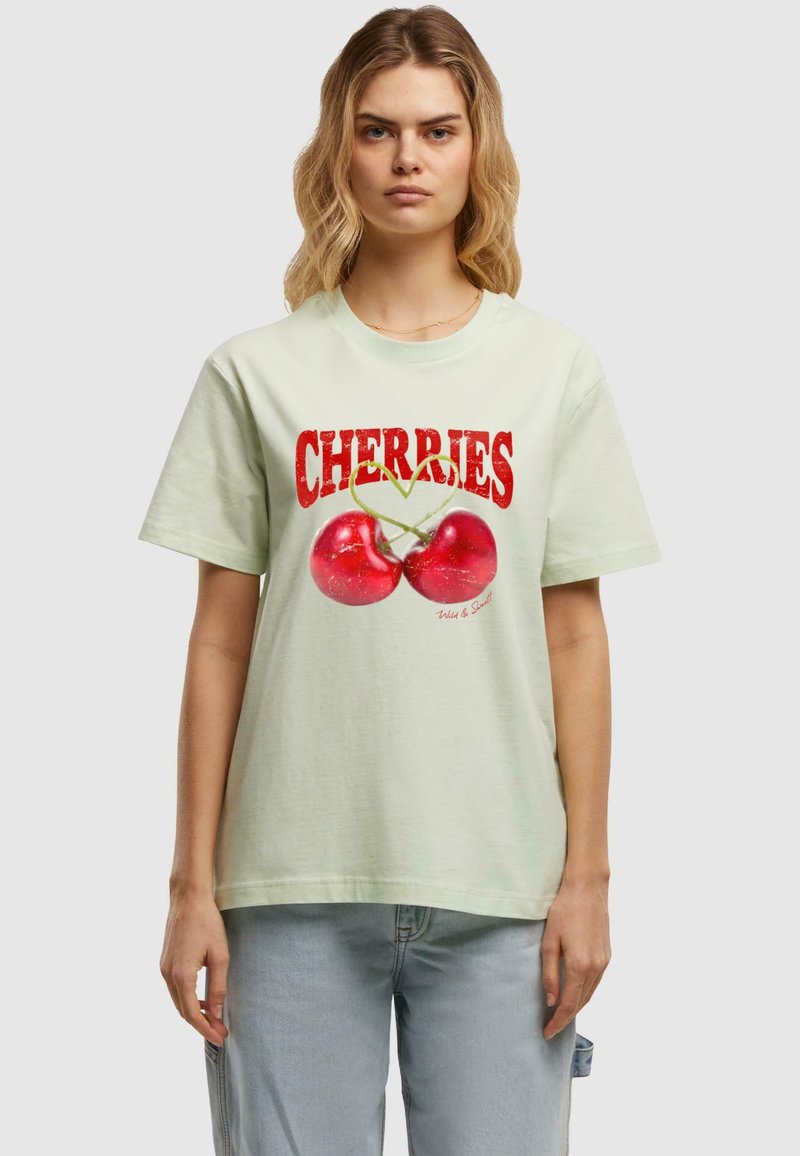 Lysegrøn t-shirt med et grafik af to røde kirsebær med grønne stilke, skrift i fed rød, der siger "CHERRIES" ovenfor. Korte ærmer, rund halsudskæring.