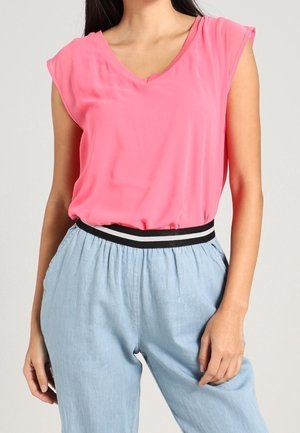 Kvinde iført en lyserød ærmeløs V-hals top, der er stoppet ned i lyseblå bukser med en sort og hvid elastisk linning.