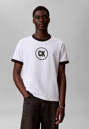 Ung mann med mørk hud som har på seg hvit T-skjorte med svart "CK"-logo i en sirkel, svart krage og ermekant, samt mørke dongeribukser, stående med den ene hånden i lomma.