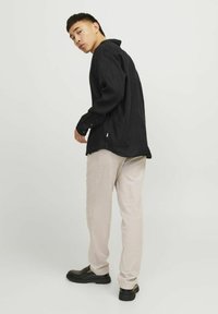 Camicia nera a maniche lunghe con colletto e polsini abbottonati, abbinata a pantaloni beige chiaro e scarpe nere senza lacci, vista da dietro.